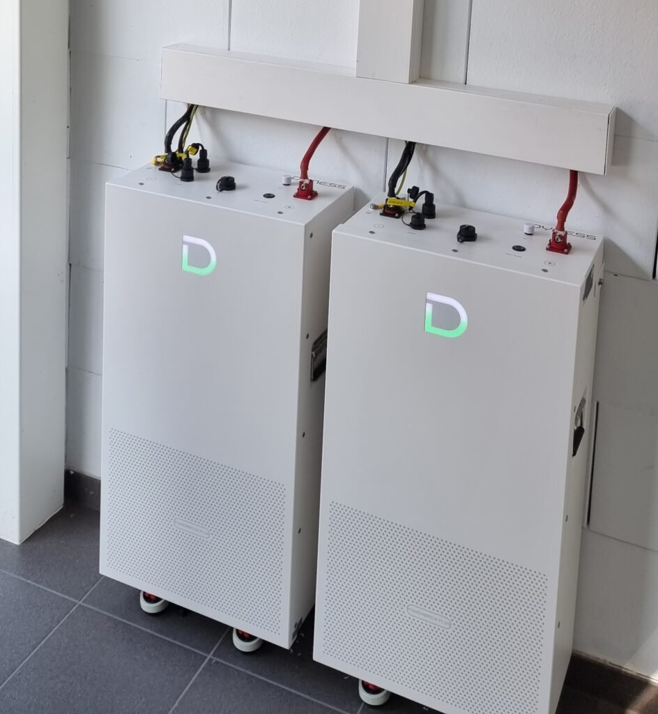 Two white battery cabinets on wheels connected with red and black cables in a utility room.